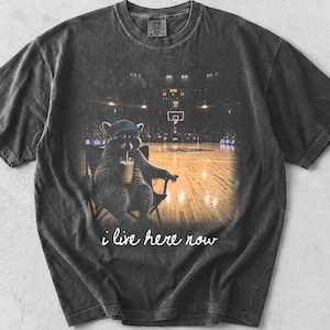 May include: Dark gray t-shirt featuring a raccoon wearing a blue cap, sitting in a chair, and drinking from a cup. The raccoon is in a basketball arena. The text "i live here now" is written in white.