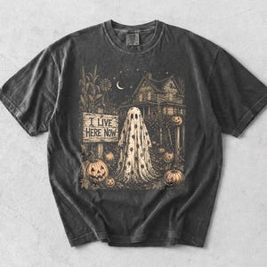 Może przedstawiać: Ciemnoszary t-shirt z krótkim rękawem z nadrukiem przedstawiającym ducha stojącego przed domem w otoczeniu dyń, łodyg kukurydzy i półksiężyca. Napis "I LIVE HERE NOW" znajduje się na drewnianej tabliczce obok postaci ducha.