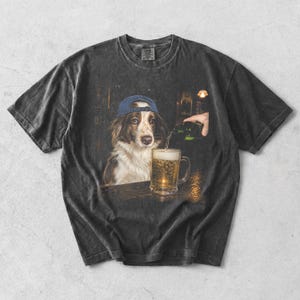 May include: A charcoal grey short-sleeve t-shirt featuring a graphic print of a brown and white dog wearing a blue baseball cap. The dog sits at a wooden bar while a hand pours a green bottle of beer into a glass mug filled with golden liquid.