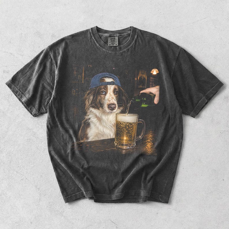 May include: A charcoal grey short-sleeve t-shirt featuring a graphic print of a brown and white dog wearing a blue baseball cap. The dog sits at a wooden bar while a hand pours a green bottle of beer into a glass mug filled with golden liquid.