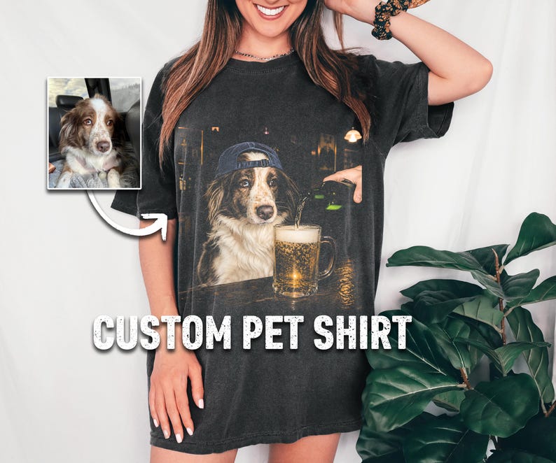 May include: A charcoal gray t-shirt featuring a graphic print of a dog wearing a blue baseball cap sitting at a bar next to a glass of beer. A small inset image shows the original dog photo. The text CUSTOM PET SHIRT is printed below.