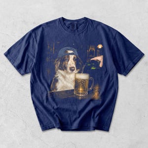 May include: A navy blue t-shirt featuring a graphic print of a brown and white dog wearing a blue baseball cap. The dog sits at a bar while a hand pours a green bottle of beer into a glass mug filled with golden liquid and white foam.