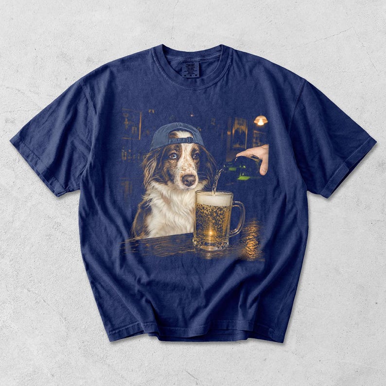 May include: A navy blue t-shirt featuring a graphic print of a brown and white dog wearing a blue baseball cap. The dog sits at a bar while a hand pours a green bottle of beer into a glass mug filled with golden liquid and white foam.