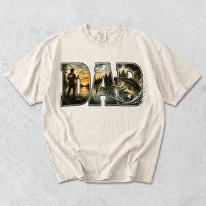 May include: Cream-colored t-shirt featuring the word "DAD" in a nature-themed design. The "D" shows a person fishing at sunset, and the "A" and "D" show a lake scene with a fish jumping out of the water. The shirt is made of a soft material.
