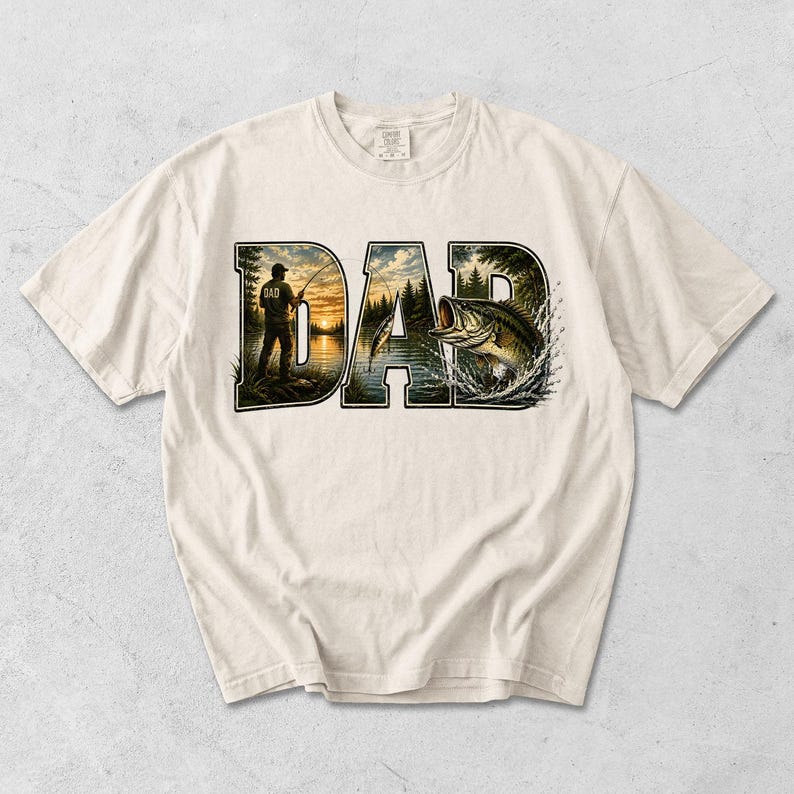 May include: Cream-colored t-shirt featuring the word "DAD" in a nature-themed design. The "D" shows a person fishing at sunset, and the "A" and "D" show a lake scene with a fish jumping out of the water. The shirt is made of a soft material.