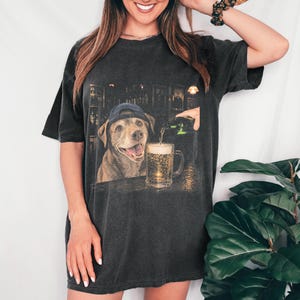 May include: A charcoal grey short-sleeve t-shirt featuring a graphic print of a brown dog wearing a blue baseball cap. The dog is positioned at a bar counter next to a glass mug being filled with beer from a green bottle in a dimly lit setting.