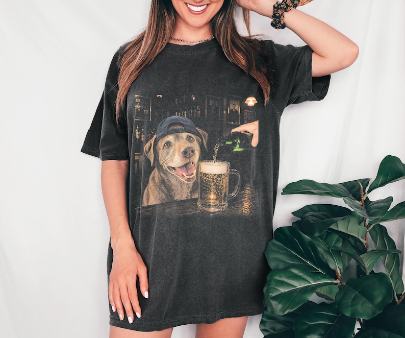 May include: A charcoal grey short-sleeve t-shirt featuring a graphic print of a brown dog wearing a blue baseball cap. The dog is positioned at a bar counter next to a glass mug being filled with beer from a green bottle in a dimly lit setting.