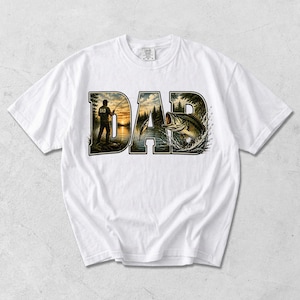 May include: White t-shirt with the word "DAD" in a stylized design featuring a fishing scene. The letters are filled with a sunset, trees, a fisherman, and a leaping fish, perfect for a fishing enthusiast.