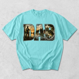 May include: A light blue t-shirt featuring the word "DAD" in a stylized design. The letters incorporate a fishing scene with a person fishing and a leaping fish. The shirt is a comfortable, casual wear item.
