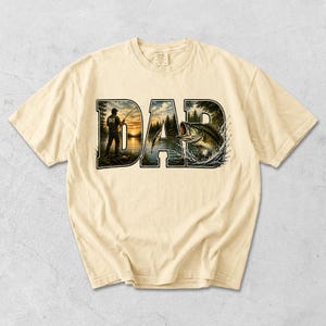 May include: Cream-colored t-shirt featuring the word "DAD" in a bold, stylized font. The letters are filled with a fishing scene, including a fisherman, a lake, and a leaping fish. The shirt has short sleeves and a crew neck.