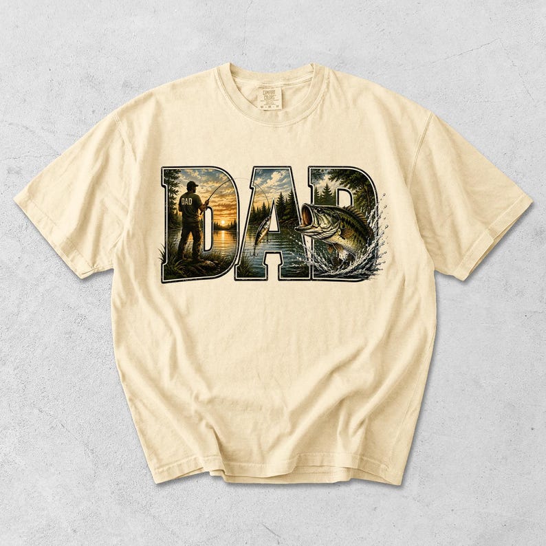 May include: Cream-colored t-shirt featuring the word "DAD" in a bold, stylized font. The letters are filled with a fishing scene, including a fisherman, a lake, and a leaping fish. The shirt has short sleeves and a crew neck.