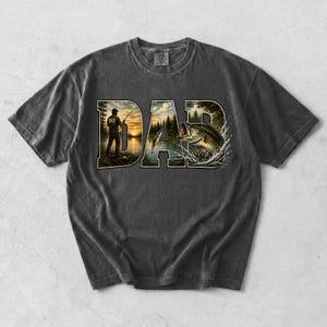 May include: Dark gray t-shirt with a graphic design featuring the word "DAD" in a nature-themed font. The letters are filled with images of a fisherman, a lake, and a leaping fish. The shirt has a vintage, washed look.