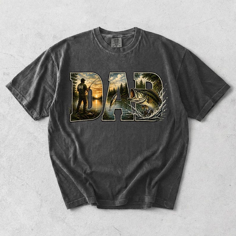 May include: Dark gray t-shirt with a graphic design featuring the word "DAD" in a nature-themed font. The letters are filled with images of a fisherman, a lake, and a leaping fish. The shirt has a vintage, washed look.
