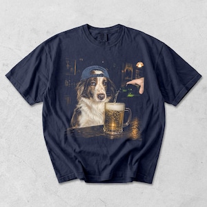 May include: A navy blue short-sleeve t-shirt featuring a graphic print of a brown and white dog wearing a blue baseball cap. The dog is positioned at a bar counter while a hand pours a green bottle of beer into a glass mug filled with golden liquid.