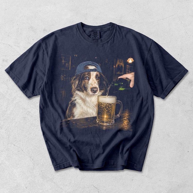 May include: A navy blue short-sleeve t-shirt featuring a graphic print of a brown and white dog wearing a blue baseball cap. The dog is positioned at a bar counter while a hand pours a green bottle of beer into a glass mug filled with golden liquid.