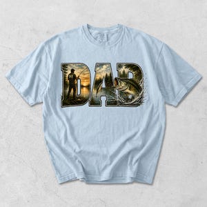 May include: Light blue t-shirt with the word "DAD" in a stylized design. The letters feature a fishing scene with a person fishing and a leaping bass. The shirt is made of a soft material.