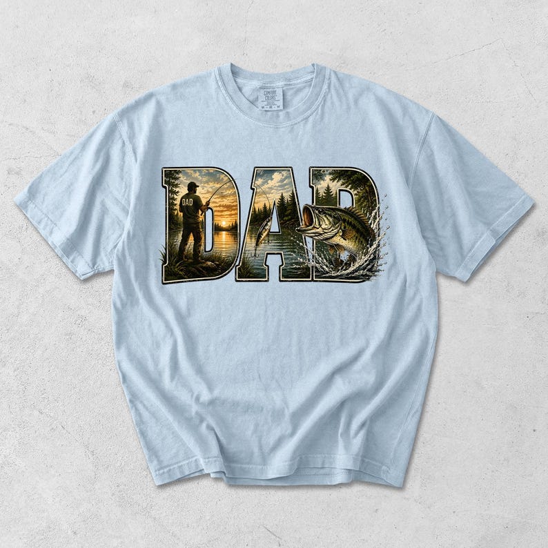 May include: Light blue t-shirt with the word "DAD" in a stylized design. The letters feature a fishing scene with a person fishing and a leaping bass. The shirt is made of a soft material.