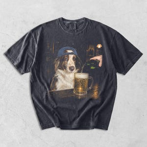 May include: A charcoal grey short-sleeve t-shirt featuring a graphic print of a brown and white dog wearing a blue baseball cap. The dog is positioned next to a glass mug being filled with beer from a green bottle in a dimly lit bar setting.