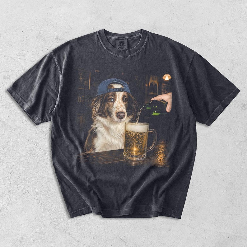 May include: A charcoal grey short-sleeve t-shirt featuring a graphic print of a brown and white dog wearing a blue baseball cap. The dog is positioned next to a glass mug being filled with beer from a green bottle in a dimly lit bar setting.