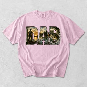 May include: Light pink t-shirt featuring the word "DAD" in a dark green, textured font. The letters are filled with a fishing scene, including a fisherman, lake, trees, and a leaping bass. The shirt is made of a soft material.