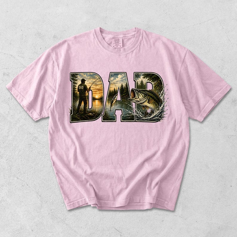 May include: Light pink t-shirt featuring the word "DAD" in a dark green, textured font. The letters are filled with a fishing scene, including a fisherman, lake, trees, and a leaping bass. The shirt is made of a soft material.