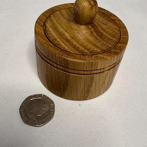 May include: A small, round wooden container with a lid and a knob on top. The container is made of light brown wood and has a circular shape. A coin is next to the container.
