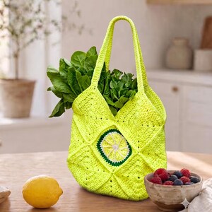 May include: A bright neon yellow crochet tote bag constructed from joined square motifs. The center of the bag features a circular lime slice design with white, light green, and dark green yarn. The bag is filled with a bunch of fresh green spinach.