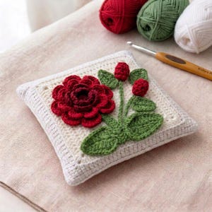 May include: A square, off-white crochet pillow featuring a large, red crochet rose with green leaves and buds. The pillow is surrounded by a white border. Red, green, and white yarn balls and a crochet hook are in the background.