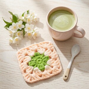 May include: A square crochet coaster featuring a light peach border and a bright green floral center, accompanied by a ceramic mug filled with a light green matcha latte, a small white spoon, and a cluster of small white jasmine flowers.