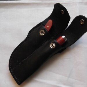 May include: A black leather sheath with two slots for holding knives. The sheath has two metal snaps and is designed to carry two knives.