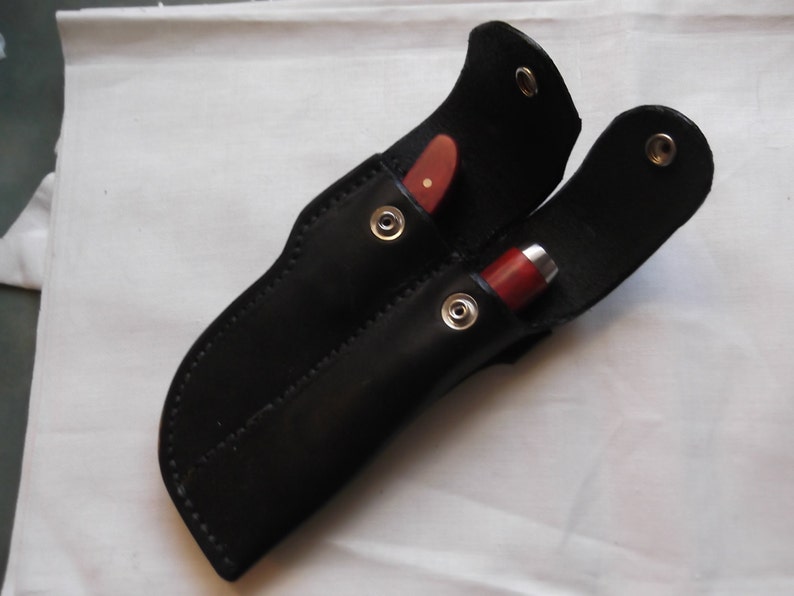 May include: A black leather sheath with two slots for holding knives. The sheath has two metal snaps and is designed to carry two knives.