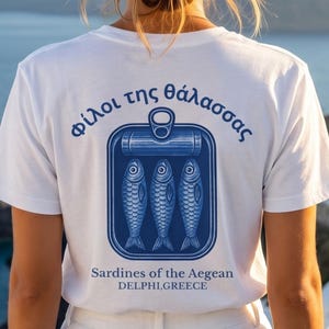 Mediterranean Greece graphic shirt featuring retro sardine can artwork and classic blue coastal print design. This vintage tinned fish tee blends European summer fashion with old world seafood packaging inspiration for a unique coastal aesthetic look. Printed on a Comfort Colors 1717 heavyweight garment dyed t shirt with relaxed fit styling, making it perfect for beach trips, seaside vacations, Greek travel lovers, foodie gifts, and vintage fashion enthusiasts.