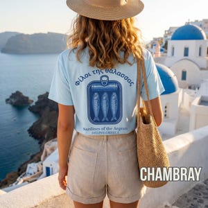 Retro fish graphic tee inspired by Greek coastal culture and Mediterranean seafood traditions. This white Comfort Colors 1717 shirt features a detailed sardine can illustration with blue nautical artwork and vintage typography printed across the back. Great for coastal aesthetic outfits, summer travel fashion, seafood lovers, tinned fish trend fans, beach vacation wardrobes, and unique vintage inspired graphic streetwear collections.