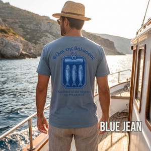 Coastal sardine shirt with vintage seafood packaging inspired artwork and Mediterranean summer style aesthetics. The oversized back print features Greek typography and retro blue sardine can graphics printed on a white Comfort Colors 1717 garment dyed t shirt. Perfect for European beach vacations, foodie fashion lovers, nautical outfit styling, retro streetwear fans, and anyone who loves unique coastal inspired apparel.