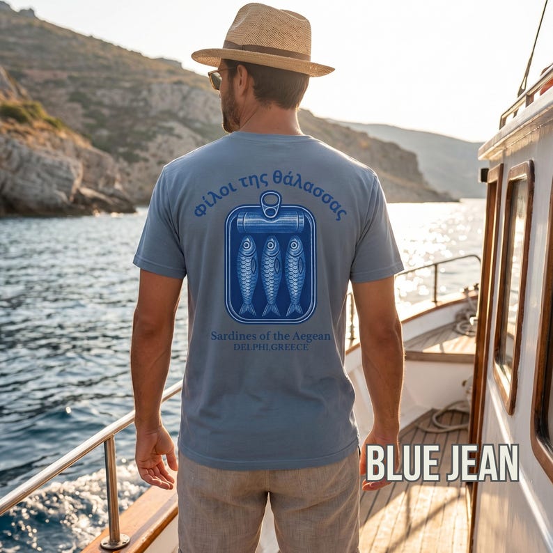 Coastal sardine shirt with vintage seafood packaging inspired artwork and Mediterranean summer style aesthetics. The oversized back print features Greek typography and retro blue sardine can graphics printed on a white Comfort Colors 1717 garment dyed t shirt. Perfect for European beach vacations, foodie fashion lovers, nautical outfit styling, retro streetwear fans, and anyone who loves unique coastal inspired apparel.