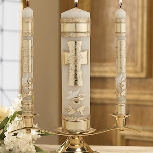 May include: A set of three white wax candles with gold metallic accents, including a large central pillar candle and two thinner taper candles, all featuring embossed cross, dove, and ring motifs, held in a polished gold metal candelabra stand.