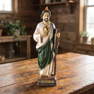 May include: A standing figurine of a bearded man wearing a cream-colored robe and a dark green cloak. He holds a brown wooden staff in one hand and a gold-colored medallion around his neck. A small flame sits atop his head. The base reads Pray for Us.