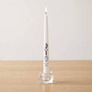 Puede incluir: Una vela cónica blanca con una llama encendida, colocada en un portavelas de cristal transparente facetado. La vela presenta un símbolo Chi-Rho de color morado oscuro, una paloma blanca, una concha de vieira y el texto "Baptized in Christ".