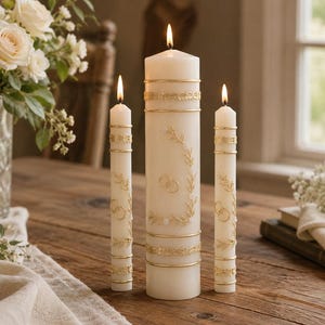 May include: A set of three off-white pillar candles with lit wicks, featuring gold-colored decorative bands and embossed gold ring and leaf motifs. The central candle is thicker than the two flanking taper candles, all standing upright.