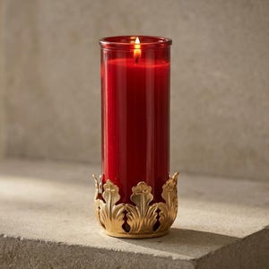 May include: A tall, cylindrical red wax candle burning inside a clear red glass vessel. The candle is held within a decorative gold-toned metal base featuring a repeating leaf-like pattern that encircles the bottom of the glass container.