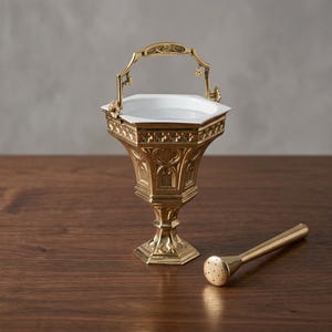 May include: A gold-toned metal holy water bucket with a white interior, featuring an ornate hexagonal base and a swinging handle, accompanied by a matching gold-toned metal sprinkler tool with a perforated head for dispensing liquid.