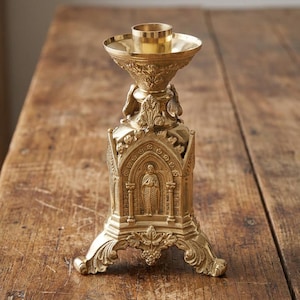 May include: A polished brass candlestick holder featuring an intricate architectural design with pointed arches, columns, and a central relief figure. The base is supported by three ornate feet decorated with leaf and grape cluster motifs.