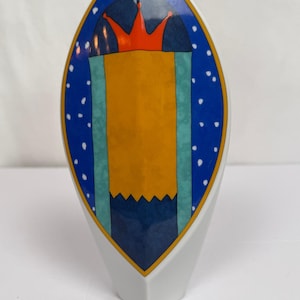 May include: A white ceramic vase with a pointed oval shape featuring a central graphic design. The design includes a bright orange crown atop a yellow rectangular panel, flanked by teal vertical stripes on a dark blue background with white dots.