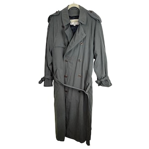 May include: A long, double-breasted trench coat in a muted gray color. The coat features a wide collar, shoulder epaulets, long sleeves with adjustable buckled cuffs, and a matching waist belt. Dark brown buttons secure the front closure.