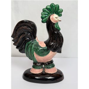 May include: A ceramic rooster figurine with a black, green, and pink color scheme. The rooster has a green crest, black feathers, and pink body. It stands on a dark brown base, showcasing a decorative and artistic design.
