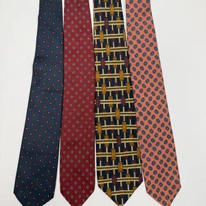 Puede incluir: Cuatro corbatas estampadas. La primera es azul marino con detalles florales rojos. La segunda es burdeos con pequeños círculos blancos y rojos. La tercera es azul marino con rectángulos dorados y burdeos. La cuarta es melocotón con estampado de cachemira azul marino.