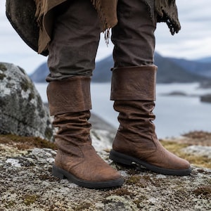 Könnte beinhalten: Braune Lederstiefel mit umgeschlagenem Schaft. Die Stiefel haben ein strukturiertes Aussehen und eine flache Sohle. Das Bild zeigt die Stiefel von den Knien abwärts, wobei die Person, die sie trägt, auf einer felsigen Oberfläche steht. Die Stiefel scheinen in einem historischen oder Fantasy-Setting getragen zu werden.