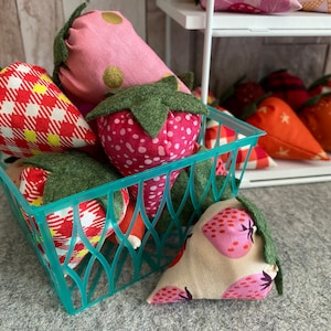 Puede incluir: Una cesta turquesa llena de adornos de fresas de tela. Las fresas son de varios tonos de rosa, rojo y beige, con hojas de tela verde. Algunas tienen lunares u otros patrones. Hay m&aacute;s fresas en una estanter&iacute;a blanca.