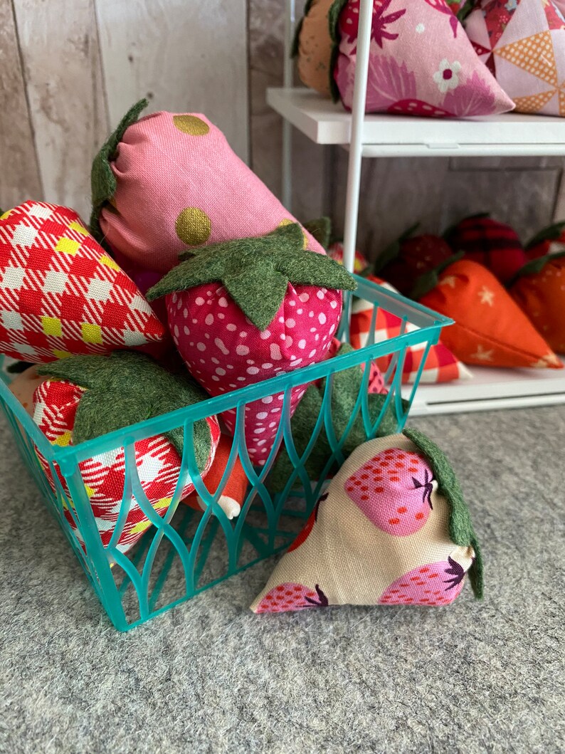 Puede incluir: Una cesta turquesa llena de adornos de fresas de tela. Las fresas son de varios tonos de rosa, rojo y beige, con hojas de tela verde. Algunas tienen lunares u otros patrones. Hay m&aacute;s fresas en una estanter&iacute;a blanca.