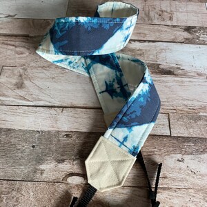 May include: A camera strap featuring a blue and white tie-dye design. The fabric strap has cream-coloured ends and black connectors. The strap is designed to be worn around the neck for ease of use.